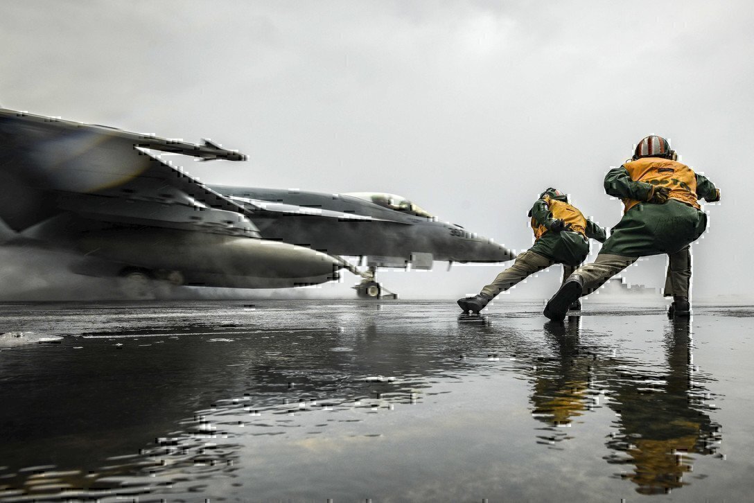 photo shows two sailors and a fighter jet taking off