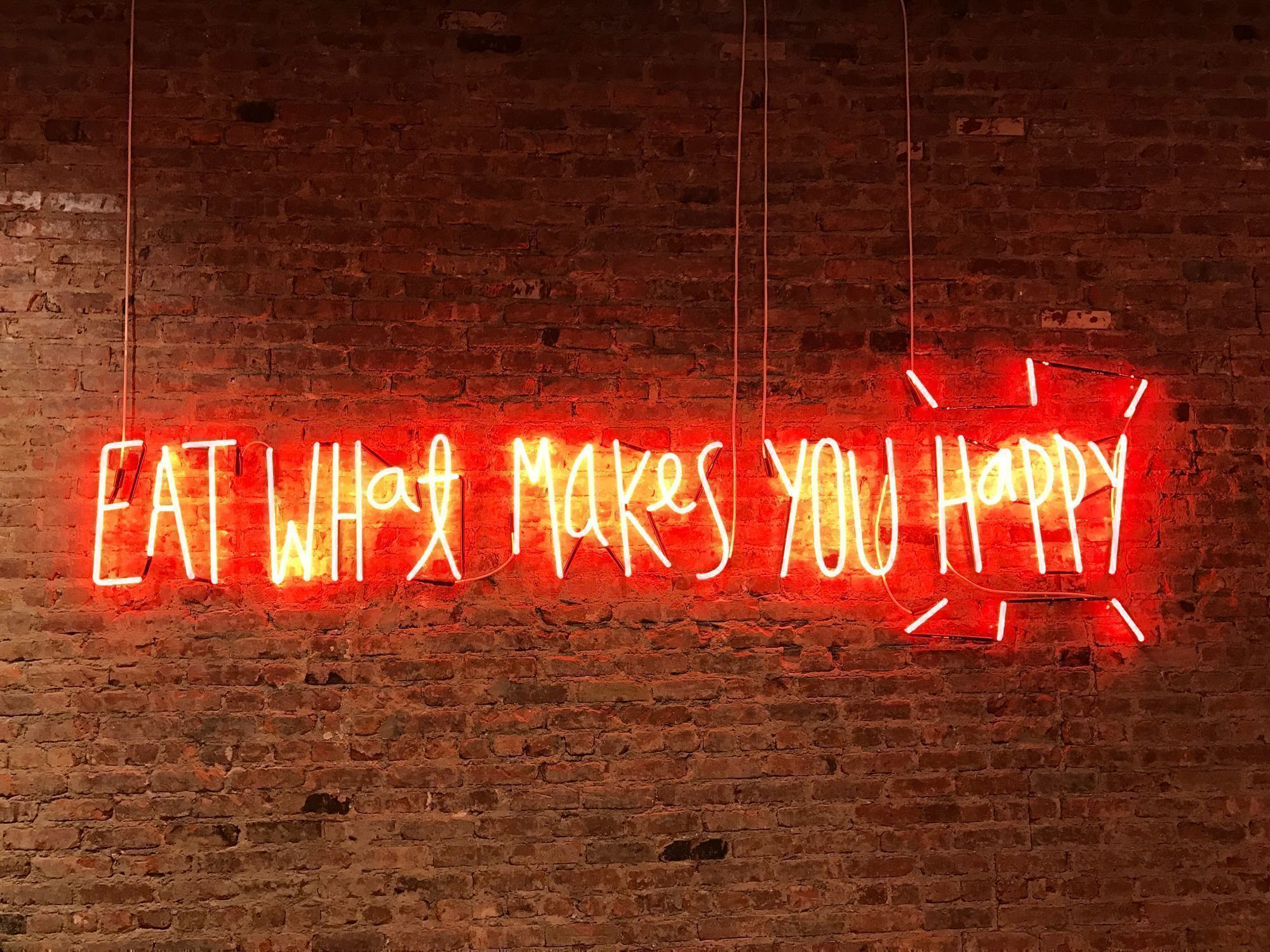 A red neon sign against a brick wall that says eat what makes you happy