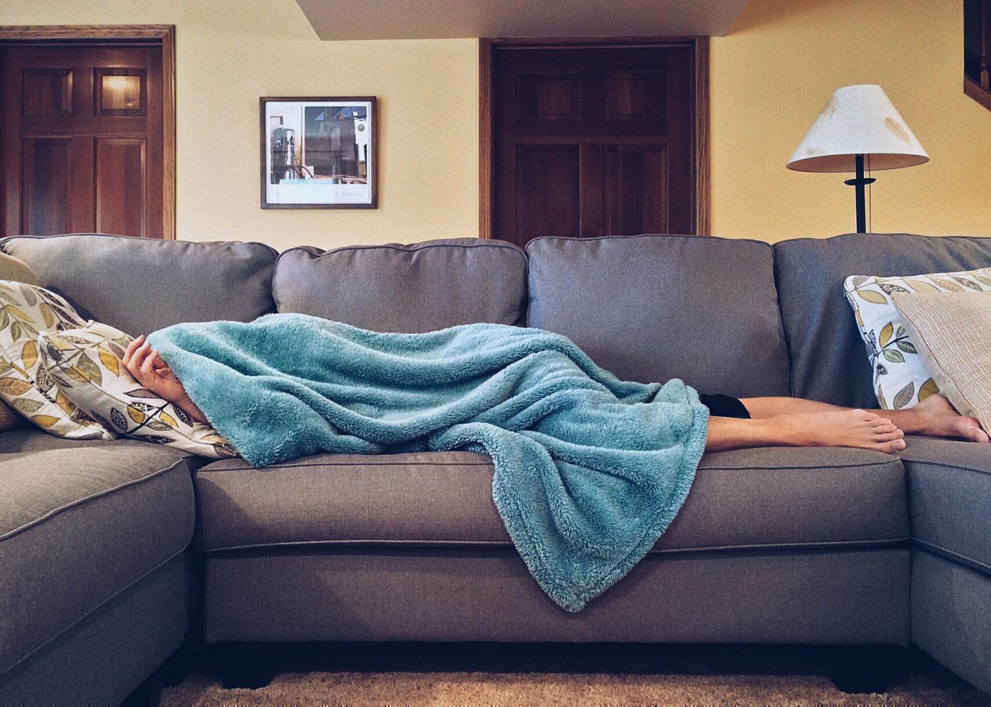 a sick person lays on a couch with a blanket over their head