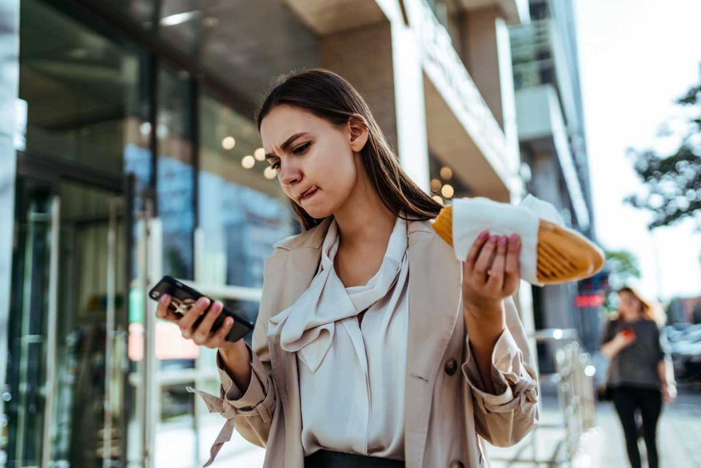 How Long Can You Take to Respond to a Job Offer?Professional woman checking her phone while eating a sandwich on the sidewalk after getting a job offer.