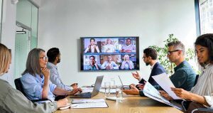 Employees on a conferece call sitting around a desk talking to remote employees on the monitor in the room.