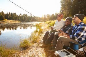 Three people sitting on the bank of a lake fishing. A little boy is sitting on his grandpa's lap watching. The younger male is holding the fishing rod and waiting.