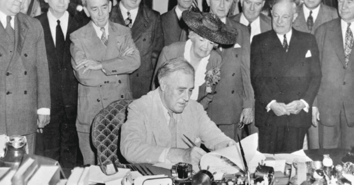 Man in a light suit signs documents at a desk as a crowd of suited colleagues watches in a formal office setting.