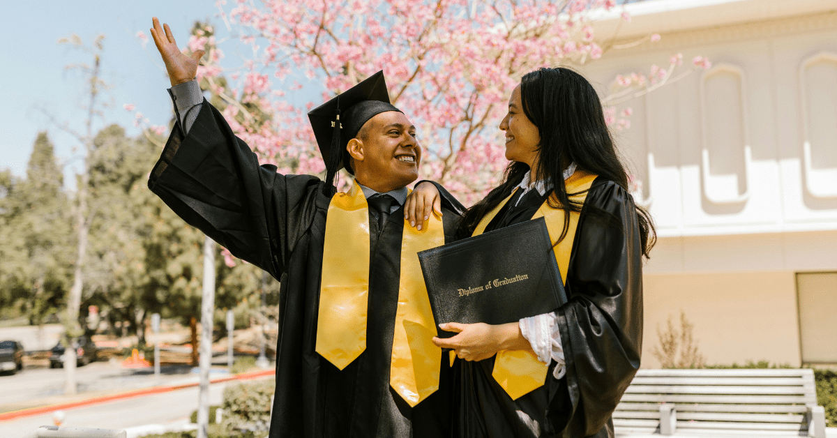 Two graduates in black gowns and gold stoles celebrate outdoors, one waving while the other holds a diploma.