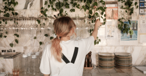 Back view of a person in a white shirt and backpack reaching toward hanging plants above a marble counter with glasses and plates nearby in a cafe setting.
